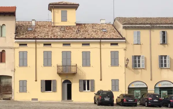 Edificio giallo con intonachino silossanico, tegole rosse, persiane grigie e balcone. Auto parcheggiate davanti.