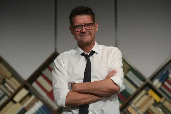 Wojciech Malajkat w białej koszuli i krawacie, z okularami na nosie, stoi na tle regałów z książkami. Jego filmy to klasyka polskiego kina.