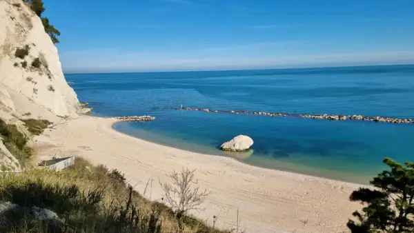 Spiaggia dorata e mare cristallino sulla Riviera del Conero, con scogliere bianche e un grande masso solitario nell'acqua.
