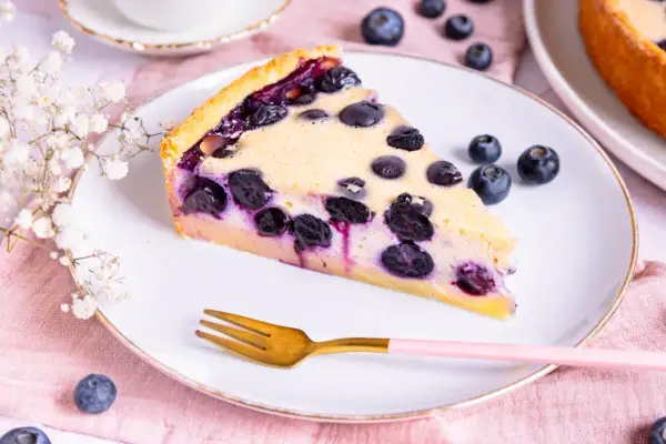 Ein Stück Heidelbeerkuchen Landfrauenart auf einem Teller, garniert mit frischen Heidelbeeren und einer kleinen Gabel.