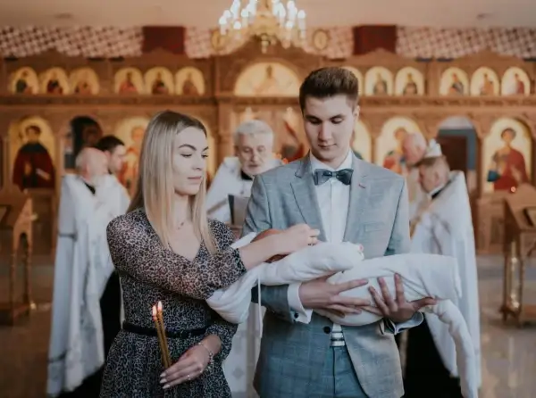 Młodzi rodzice trzymają dziecko do chrztu prawosławnego. W tle widać cerkiew i duchownych.
