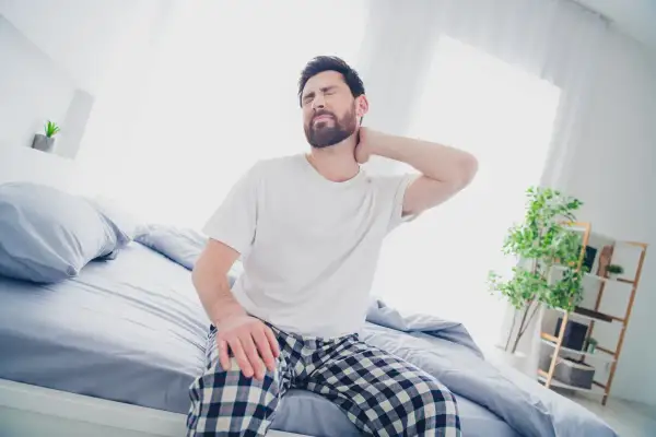 Uomo con torcicollo dopo aver dormito, si massaggia il collo con espressione sofferente, seduto sul letto.