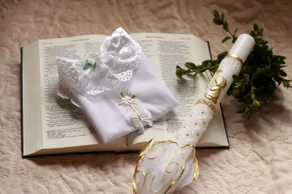 Biała szata, świeca z aniołkiem i gałązka symbol chrztu na otwartej Biblii.