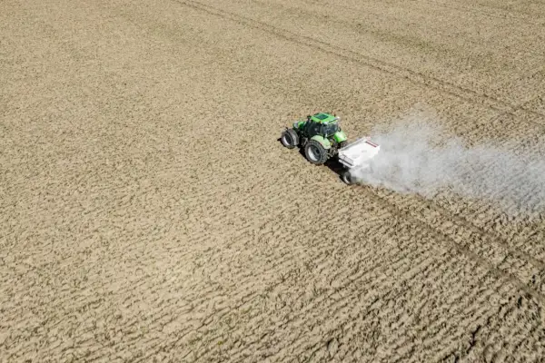 Zielony traktor z przyczepą rozsypuje nawóz na polu. To ważny etap prac polowych, który zapewnia prawidłowy wzrost roślin. Nikosar etykieta na przyczepie informuje o zawartości.