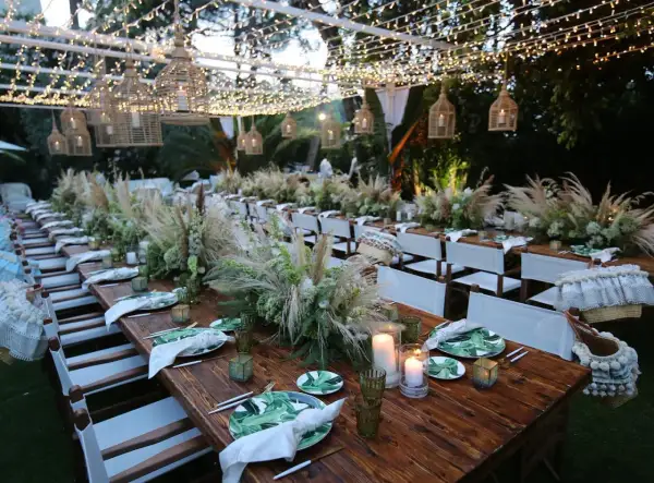 Tavoli in legno apparecchiati per un matrimonio in campagna, con centrotavola di fiori e pampas, luci soffuse e cesti decorativi.