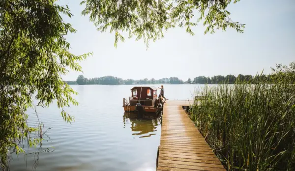 Ein Mann steht auf einem Holzsteg neben einem Hausboot auf einem See. Perfekter Urlaub in Brandenburg.