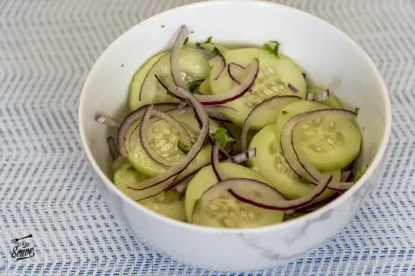 Deliciosa ensalada de pepino en rodajas finas con cebolla morada y hierbas frescas, servida en un cuenco blanco sobre un mantel azul y blanco.