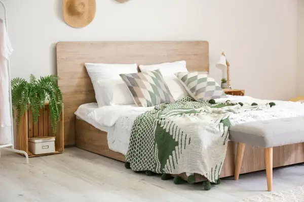 Gemütliches Schlafzimmer mit einem modernen **Aufbau Boxspringbett** in Holzoptik, grünen Decken und Kissen, sowie einer Hängepflanze.
