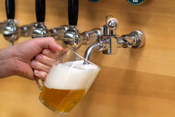 Mano che versa una birra dorata con schiuma abbondante da un rubinetto cromato. La birra alder è pronta da gustare.