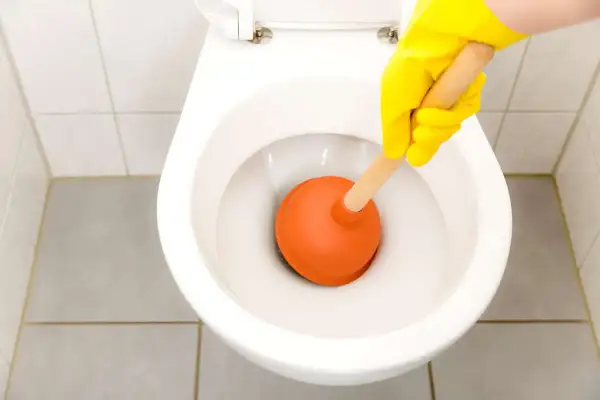 Mano con guanto giallo usa uno stantuffo arancione per liberare un filo d'acqua che scende nel water.