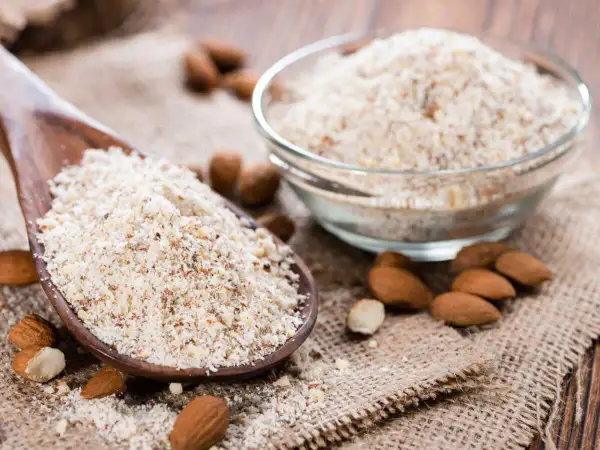 Cuillère en bois remplie de poudre d'amande, idéale pour remplacer la farine dans vos recettes. Amandes entières et bol de poudre à côté.