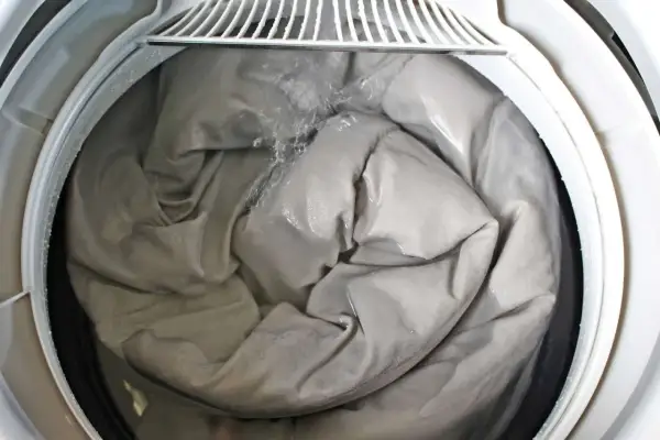 A grey comforter is being washed in a washing machine, showing the best way to wash a comforter. Water is splashing onto the fabric.
