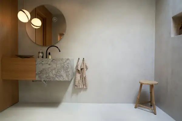 Modernt badrum med handfat i marmor och väggar i microcement. En rund spegel och två lampor ovanför.