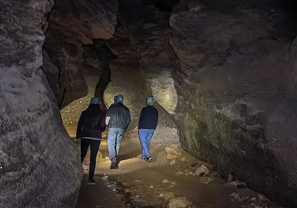 Tres personas con cascos exploran una cueva oscura, sus linternas iluminan las paredes rocosas y el suelo irregular. Los yesos naturales brillan en la oscuridad.