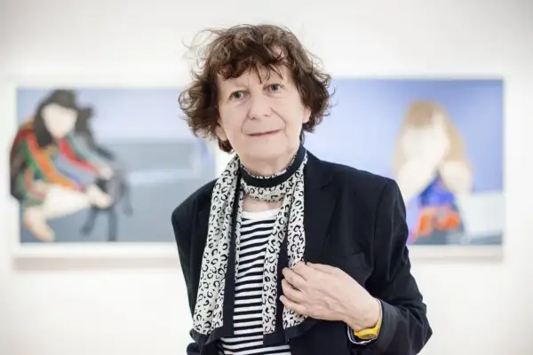 Ewa Kuryluk, artystka o burzy brązowych włosów, w czarnej marynarce i pasiastym szalu, pozuje na tle swoich obrazów.