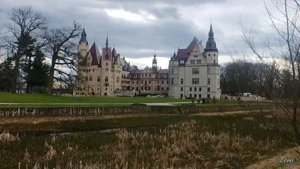 Majestatyczny zamek w Głogówku z wieżami i czerwonymi dachami, otoczony zielonym trawnikiem i stawem z trzcinami.