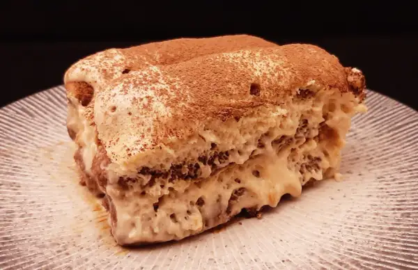 Une part de tiramisu au café, saupoudrée de cacao, sur une assiette striée. Un délice qui invite à découvrir la recette.