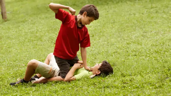 Un niño en camiseta roja agrede a otro en el suelo. Un ejemplo de cómo los niños que excluyen a otros pueden causar daño.