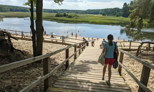 Dziewczynka schodzi drewnianymi schodami na piaszczystą plażę nad jeziorem. W oddali widać ludzi kąpiących się i odpoczywających. Stawy echo zwierzyniec to idealne miejsce na letni relaks.