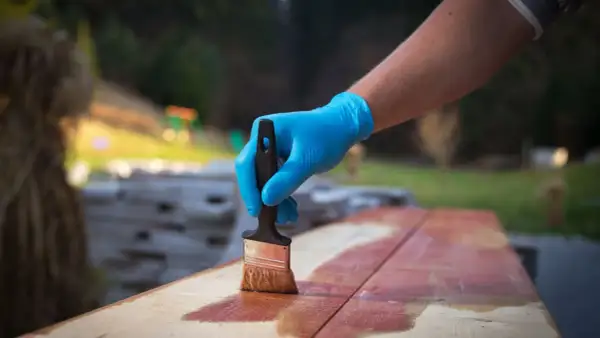 Main tenant une brosse, applique une lasure sur des planches de bois à l'extérieur.