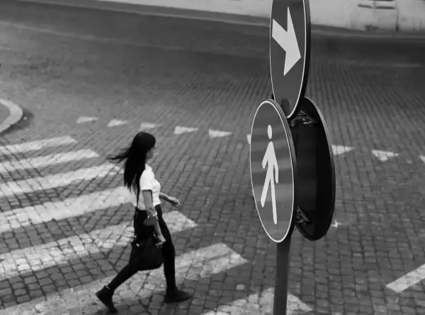 Una donna attraversa la strada su un selciato, seguendo le indicazioni di un segnale pedonale. Fotografia di strada.