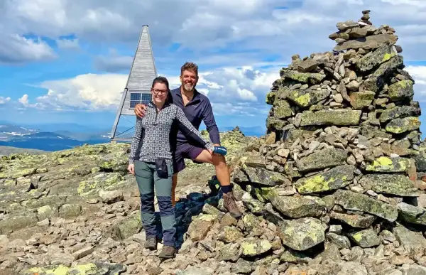 Två glada vandrare vid en stenröse på en bergstopp i Sverige. En modern, pyramidformad byggnad med solpaneler syns i bakgrunden. Perfekt för att utforska Sveriges vandringsleder.