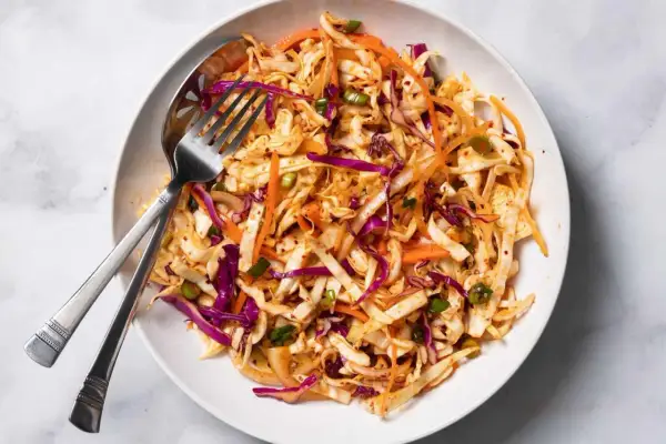 En skål med krispig kål, morötter och rödlök, perfekt för ett enkelt coleslaw recept. En gaffel och sked ligger i skålen.