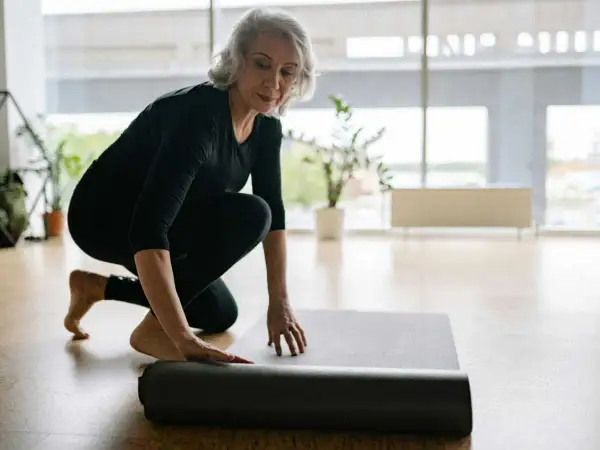 Une femme prépare son tapis pour faire du sport chez soi. Elle est à genoux, déroulant le tapis gris sur le sol en bois.
