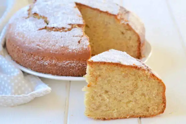 Una fetta di torta di banane soffice, cosparsa di zucchero a velo, accanto alla torta intera su un piatto bianco.