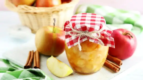 Frisch eingekochtes Apfelkompott im Glas, dekoriert mit kariertem Stoff und Zimtstangen. Äpfel im Korb im Hintergrund.