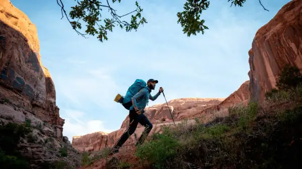 Hombre con mochila y bastones de senderismo sube una ladera rocosa bajo un cielo azul.
