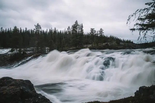 Vilt och mäktigt vattenfall i Norrland, med tät barrskog som bakgrund och en dramatisk, grå himmel.