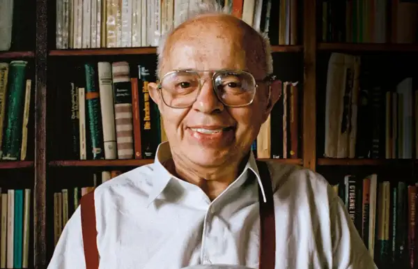 Uśmiechnięty Stanisław Lem, autor powieści Solaris, siedzi przed regałami pełnymi książek.