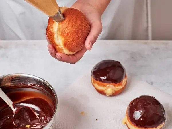 Eine Hand füllt einen Boston Cream Donut mit cremiger Füllung. Daneben liegen bereits glasierte Donuts.