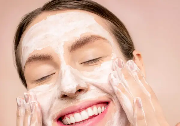 Donna sorridente con maschera bianca sul viso, mentre impara come fare una pulizia del viso a casa.