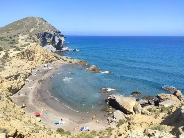 Vista aérea de **playas nudistas Cabo de Gata**. Arena oscura, aguas turquesas y formaciones rocosas. Gente disfrutando del sol y el mar.
