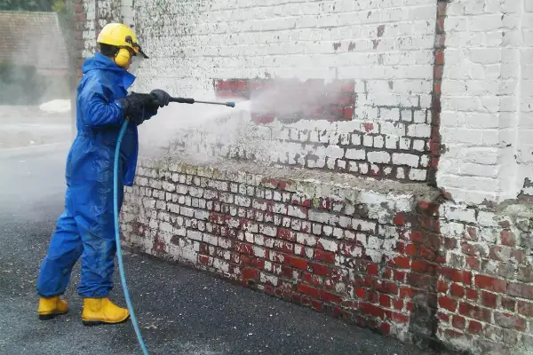 Personne en combinaison bleue et bottes jaunes utilise un nettoyeur haute pression pour enlever peinture sur brique.