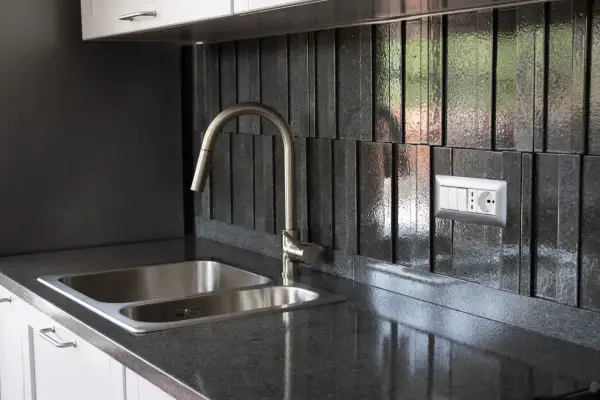 Top cucina in pietra lavica: pro e contro. Un lavello doppio in acciaio inox e un rubinetto moderno su un piano scuro e un rivestimento a listelli verticali.