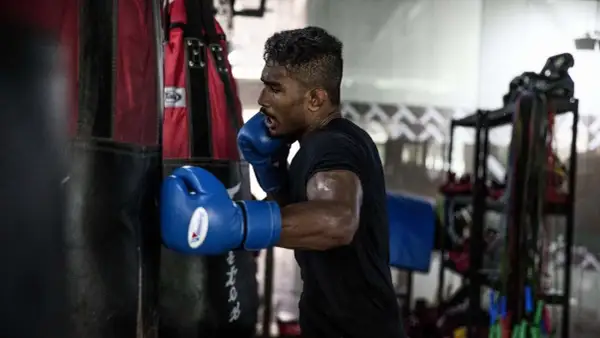 Boxer in blue gloves throwing a powerful cross punch at a heavy bag, sweat glistening on his arms.