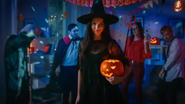 Kiedy obchodzi się Halloween, młoda kobieta w stroju czarownicy trzyma świecącą dynię na imprezie z przyjaciółmi w kostiumach.