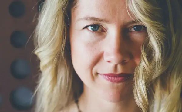 Zbliżenie na twarz kobiety z blond włosami. Delikatny uśmiech, lekko opadające policzki, niebieskie oczy.
