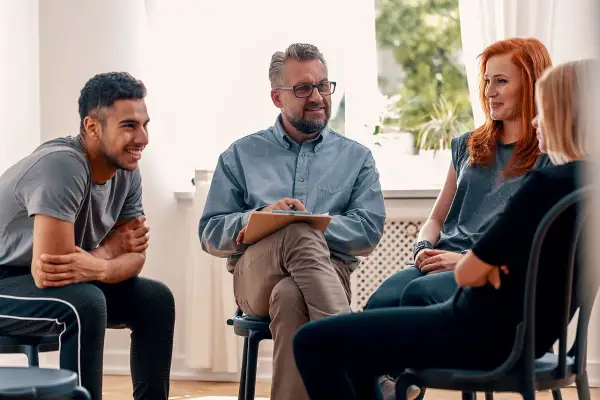 Grupa ludzi na sesji terapeutycznej. Mężczyzna z notatkami prowadzi rozmowę, a uczestnicy słuchają. Może uczą się języka francuskiego?