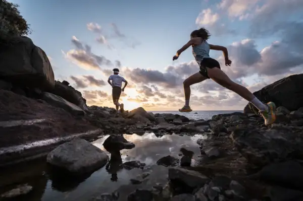 Deux coureurs s'entraînent sur un sentier rocailleux à Tenerife au coucher du soleil, l'océan en arrière-plan.