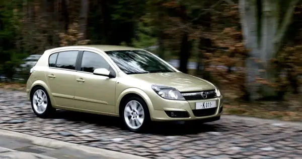 Złoty Opel Astra H 1.7 CDTI w ruchu na brukowanej drodze, otoczony jesiennym lasem.
