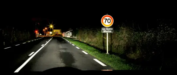 Route de nuit avec marquage au sol blanc lumineux. Panneau "Les Quatre Moulins" et limite de vitesse à 70.