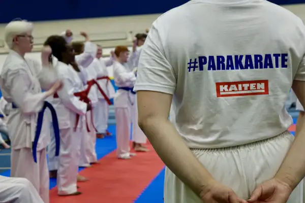En grupp utövare i vita dräkter tränar karate. En person bär en t-shirt med texten #PARAKARATE och KAITEN.