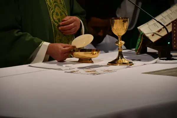 Kapłan trzyma hostię nad złotą pateną. Obok kielich i księga liturgiczna. To obrazuje przyczyny zmian w tekstach modlitw w nowym mszale rzymskim.