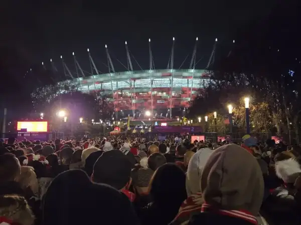 Tłum ludzi zmierza na roztanczony PGE Narodowy. Stadion oświetlony na czerwono, nocna atmosfera.