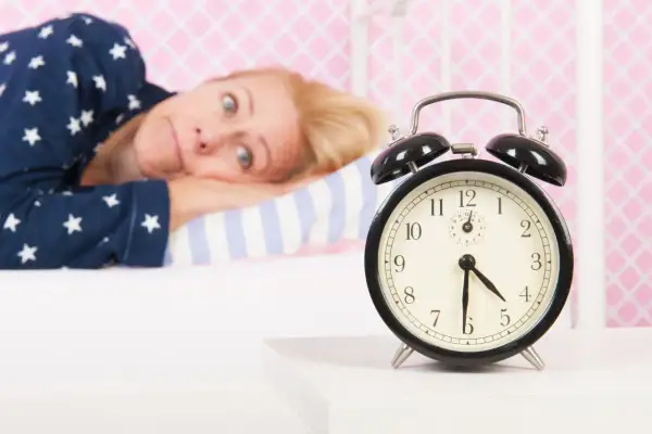 Mujer en pijama mira un despertador, su expresión refleja la **intención paradójica** de querer dormir pero no poder.