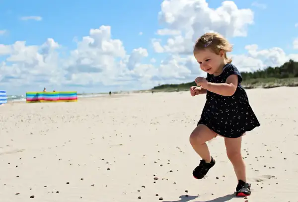 Radosna wycieczka dla dzieci na plażę. Mała dziewczynka w letniej sukience biega po piasku, ciesząc się słońcem i chmurami.